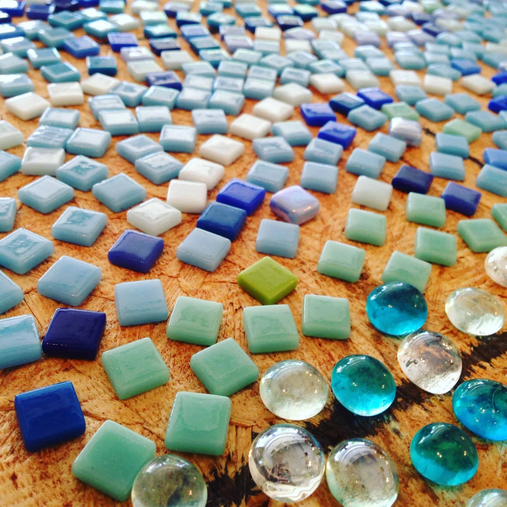 A close-up of a mosaic art piece featuring an assortment of blue, white, and green tiles alongside clear and blue round glass beads, all set on a wooden surface with the best business books stacked beside