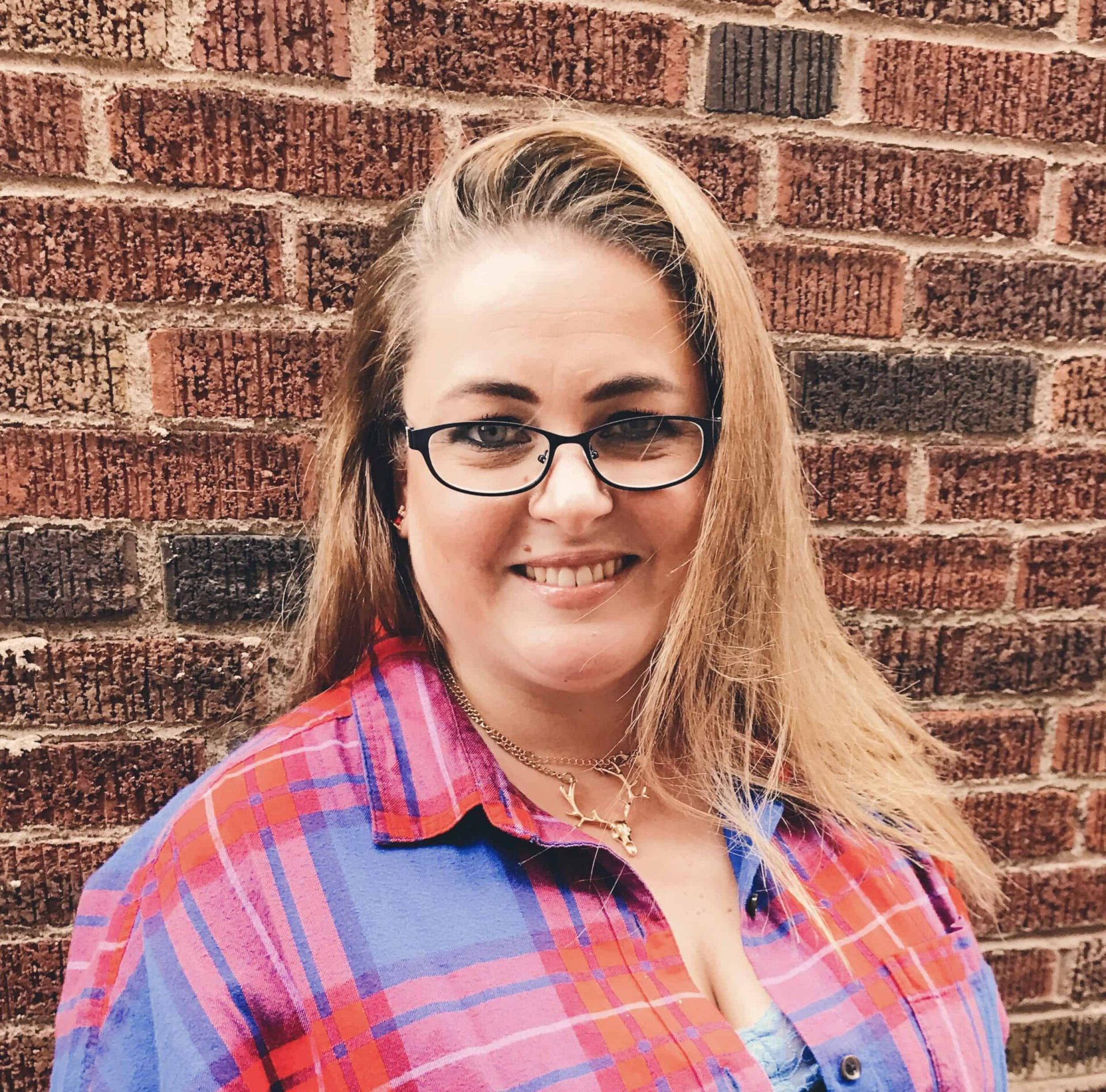 A smiling woman with shoulder-length blonde hair wearing glasses and a colorful plaid shirt stands in front of a brick wall, holding a folder labeled "Marketing Strategy.