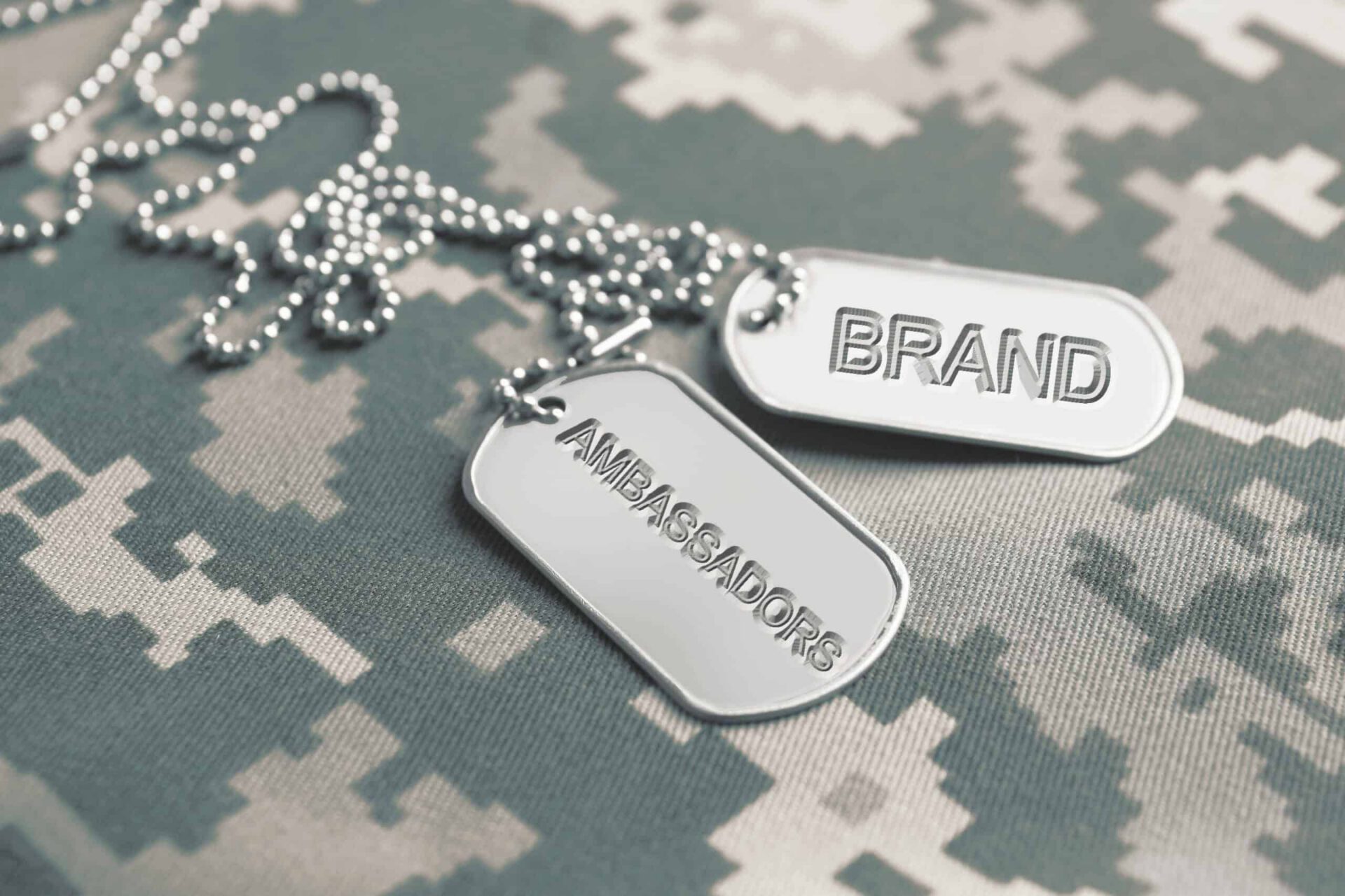 Two dog tags resting on a camouflage fabric, with one tag engraved with "brand ambassador" and the other with "ambassadors".