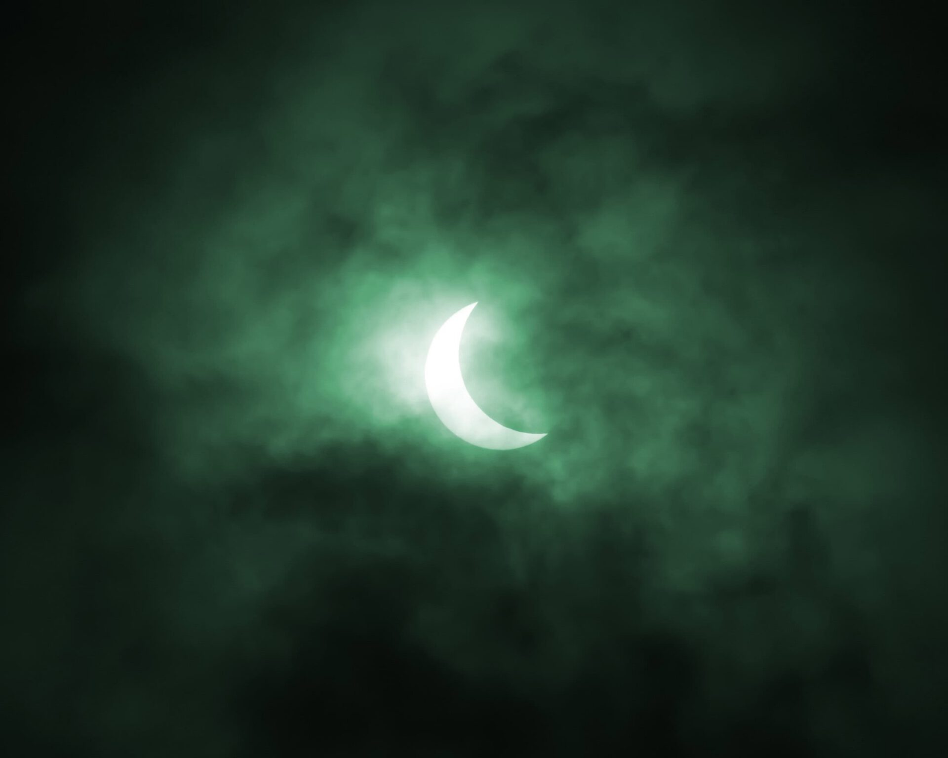 A solar eclipse viewed through a dense, green-tinted mist, showing the moon partially covering the sun, creating a crescent shape reminiscent of a brand ambassador's emblem.
