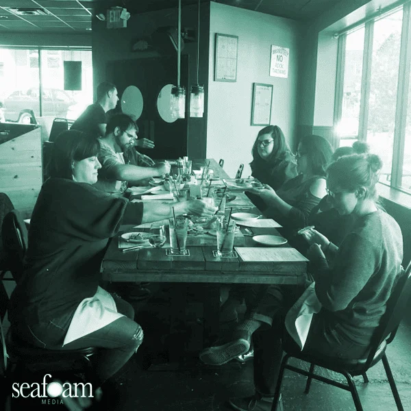 A group of people, thankful for the opportunity to dine together, are seated at a long table beside large windows in a restaurant, mostly engaged with their smartphones. The image has a monochromatic,
