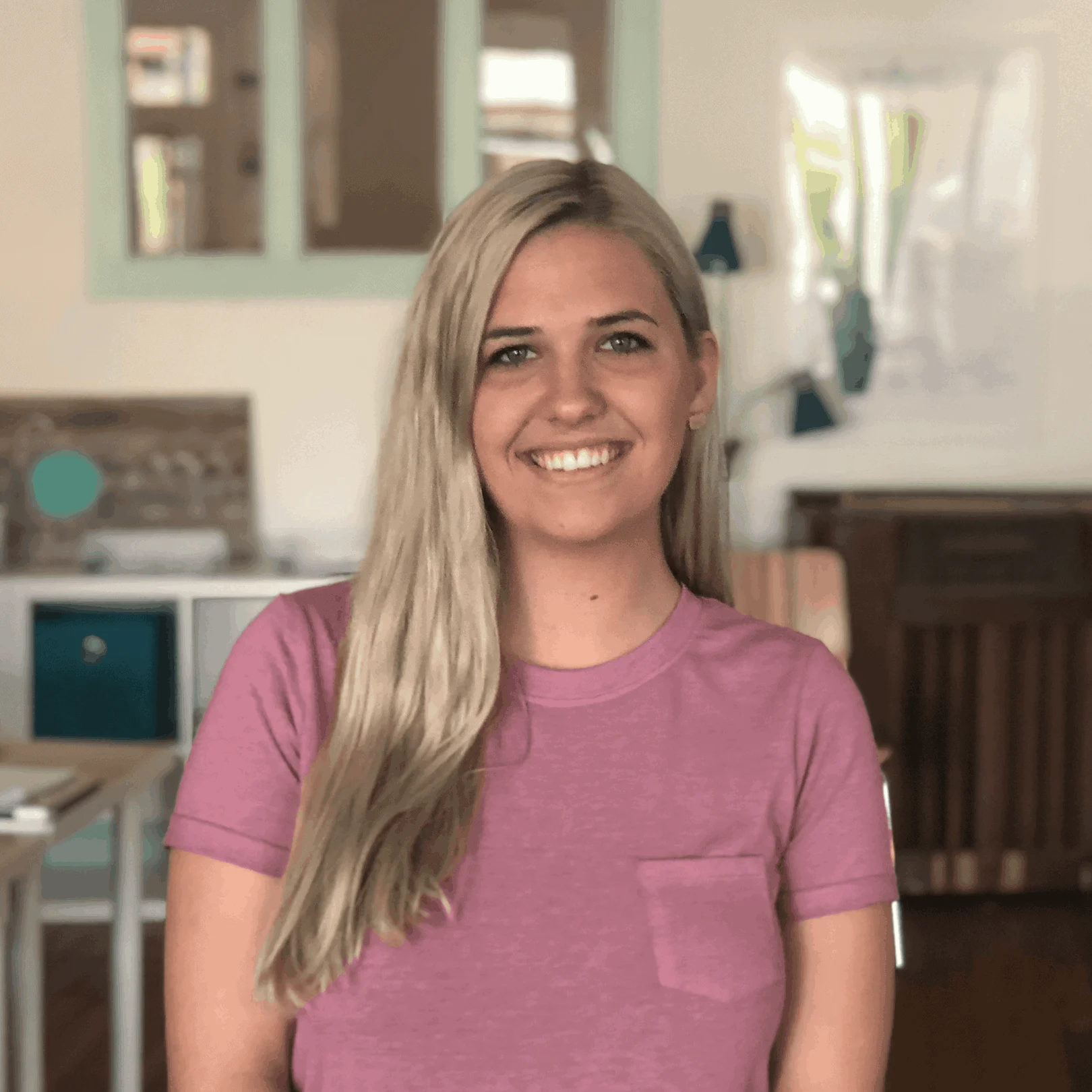 A smiling young woman with long blonde hair, wearing a purple t-shirt, stands in a room with teal and white decor as a Brand Engagement Specialist.