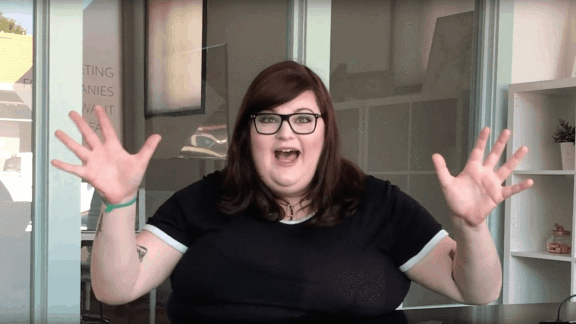 A woman with shoulder-length dark hair and glasses, wearing a black t-shirt that reads "What's Your Seashore?", excitedly talks with her hands raised, sitting in front of a bookshelf