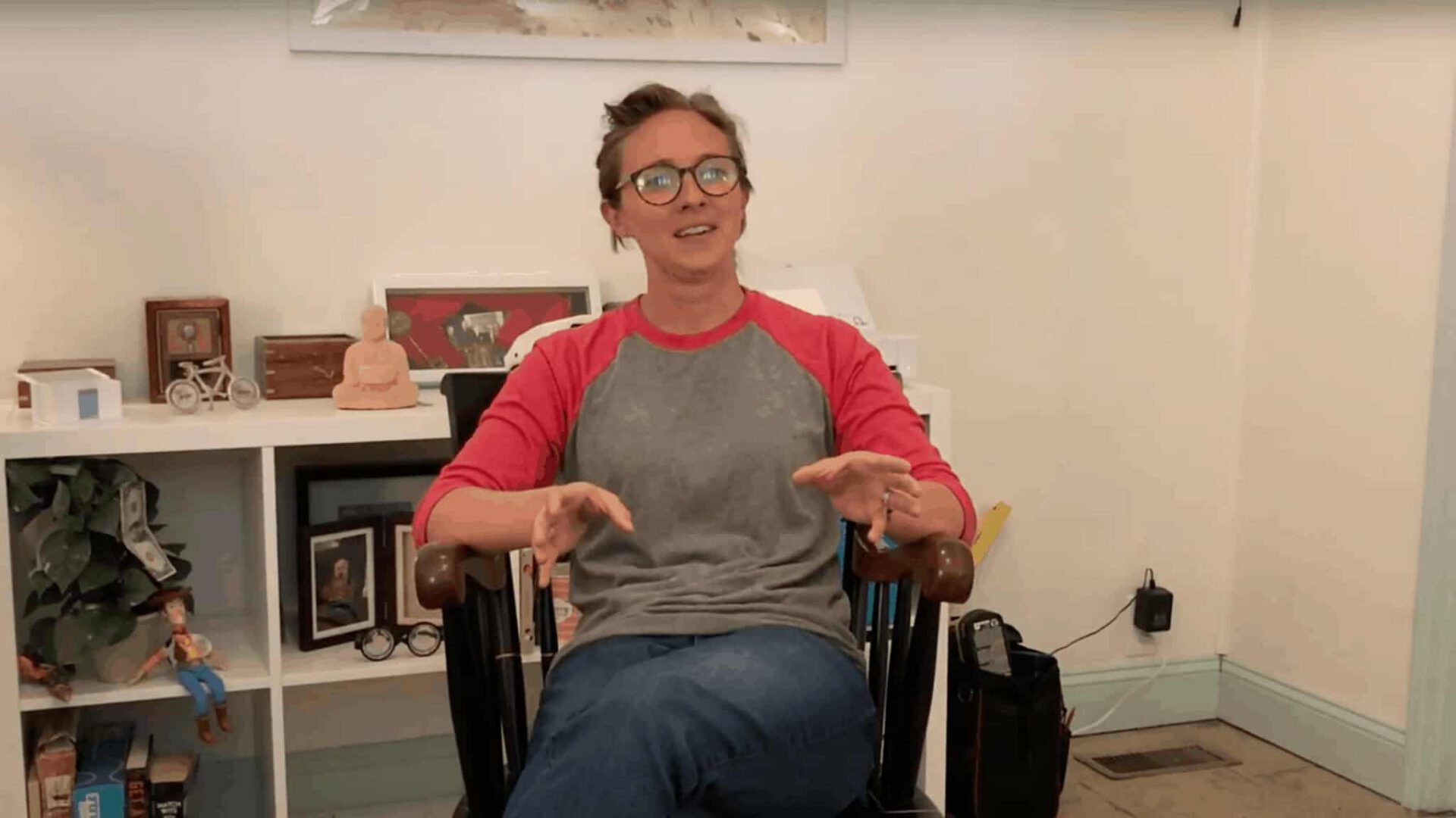 A person with glasses, wearing a gray and red shirt, sits in a wheelchair, gesturing with their hands, amidst a room filled with various decorative items and photos on shelves. "What's your