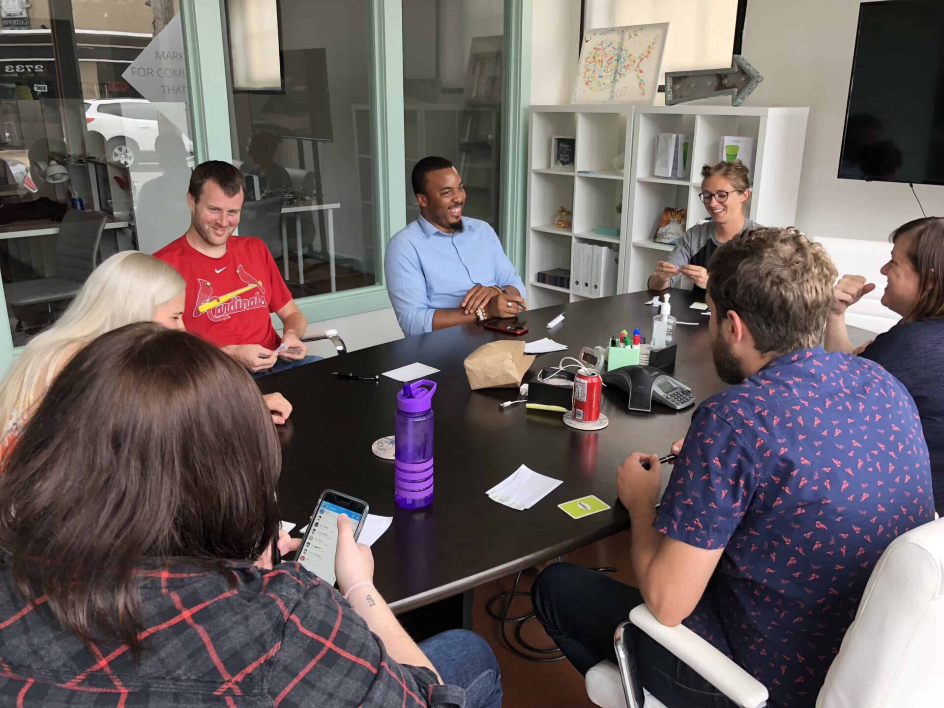 A diverse group of eight colleagues laughing and engaging in a casual meeting around a seafoam-colored table in a modern office space.