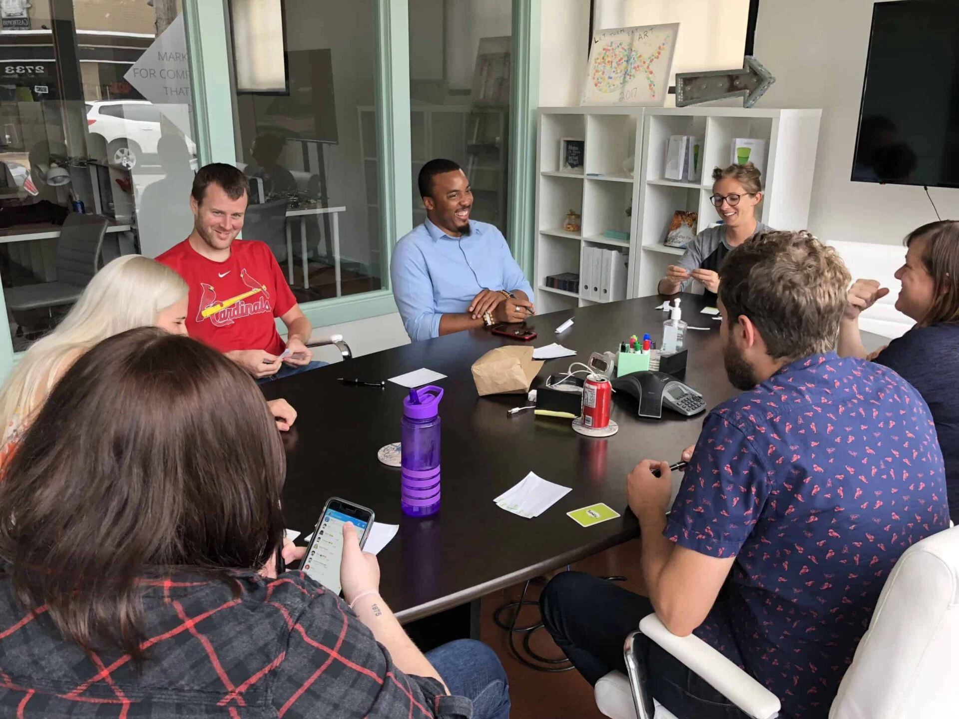 A diverse group of eight colleagues laughing and engaging in a casual meeting around a seafoam-colored table in a modern office space.
