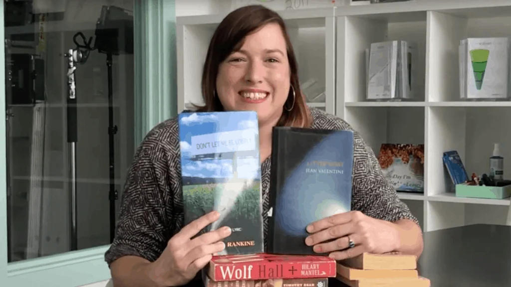 A woman smiling broadly, holding two books in front of a bookshelf filled with various books. She looks happy and enthusiastic as she ponders, "What's your seashore?