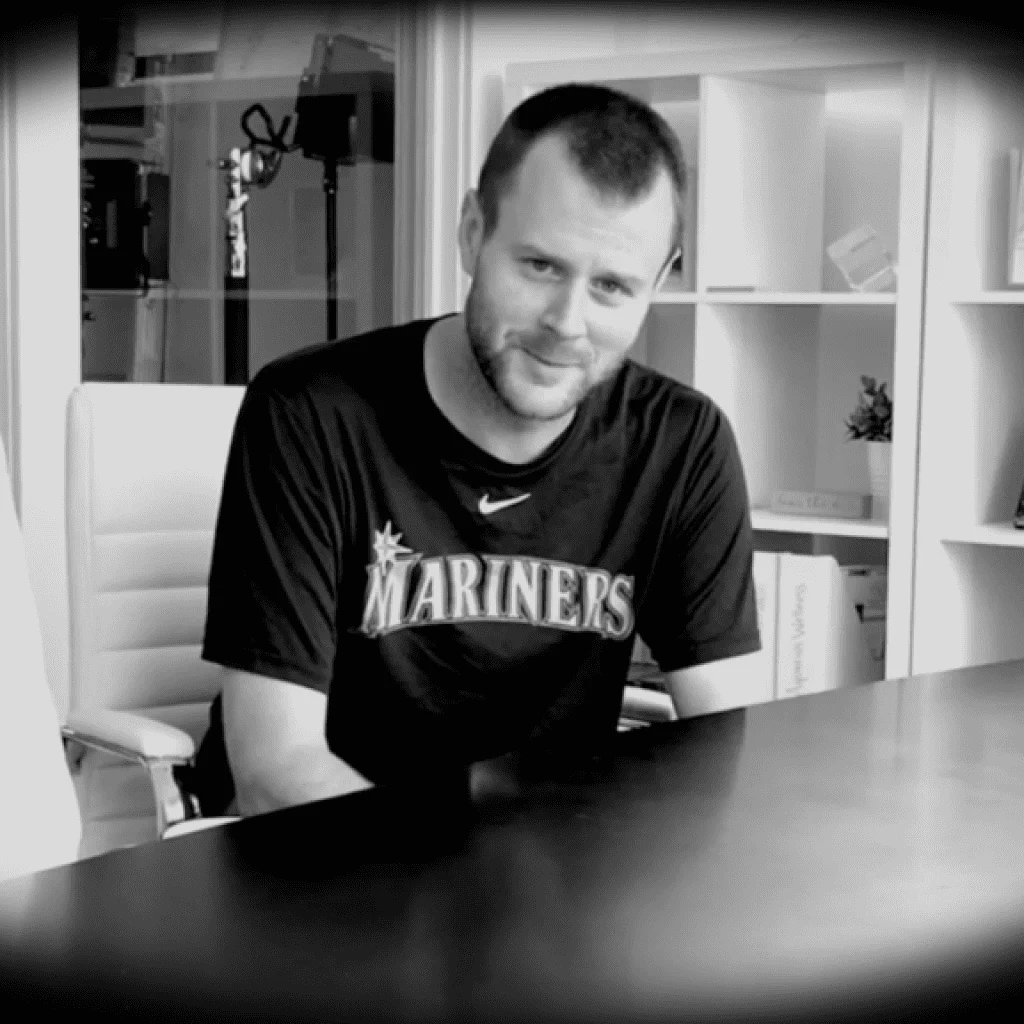 A man wearing a "What’s your Seashore?" t-shirt sits at a desk, smiling at the camera in a monochrome setting with shelves in the background.