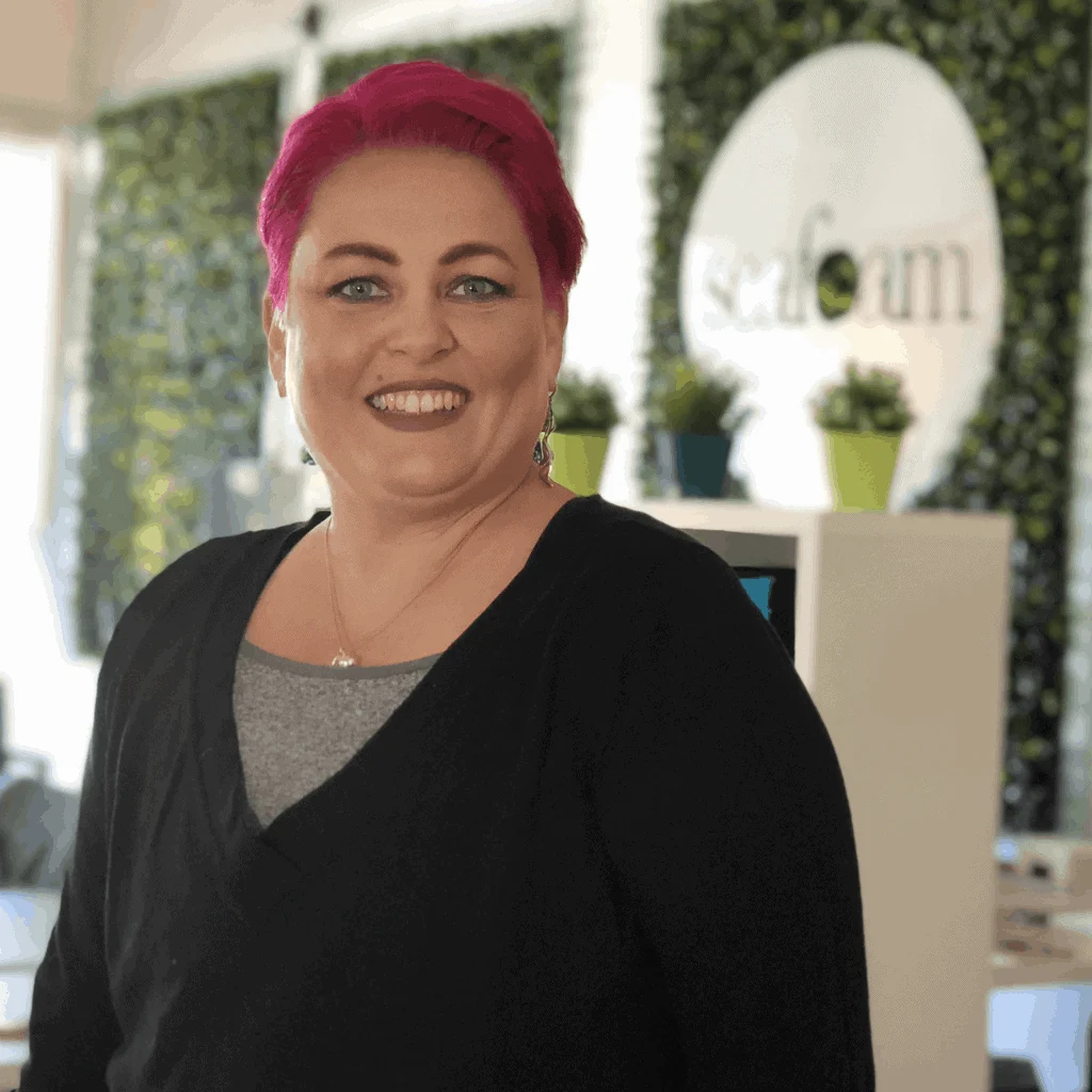 A smiling woman with vibrant pink hair standing in an office environment, with a blurred "What's Your Seashore" logo in the background and green plants around. She wears a black cardigan over