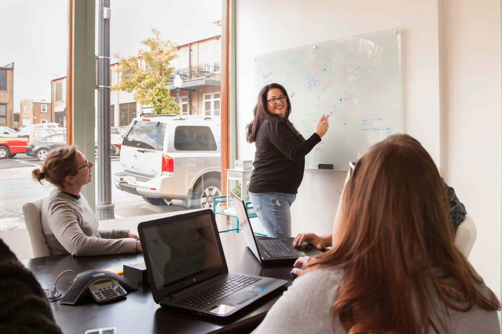 A professional woman stands presenting her New Year’s Resolutions at a whiteboard in a modern office, engaging with seated colleagues who are looking at her and laptops.