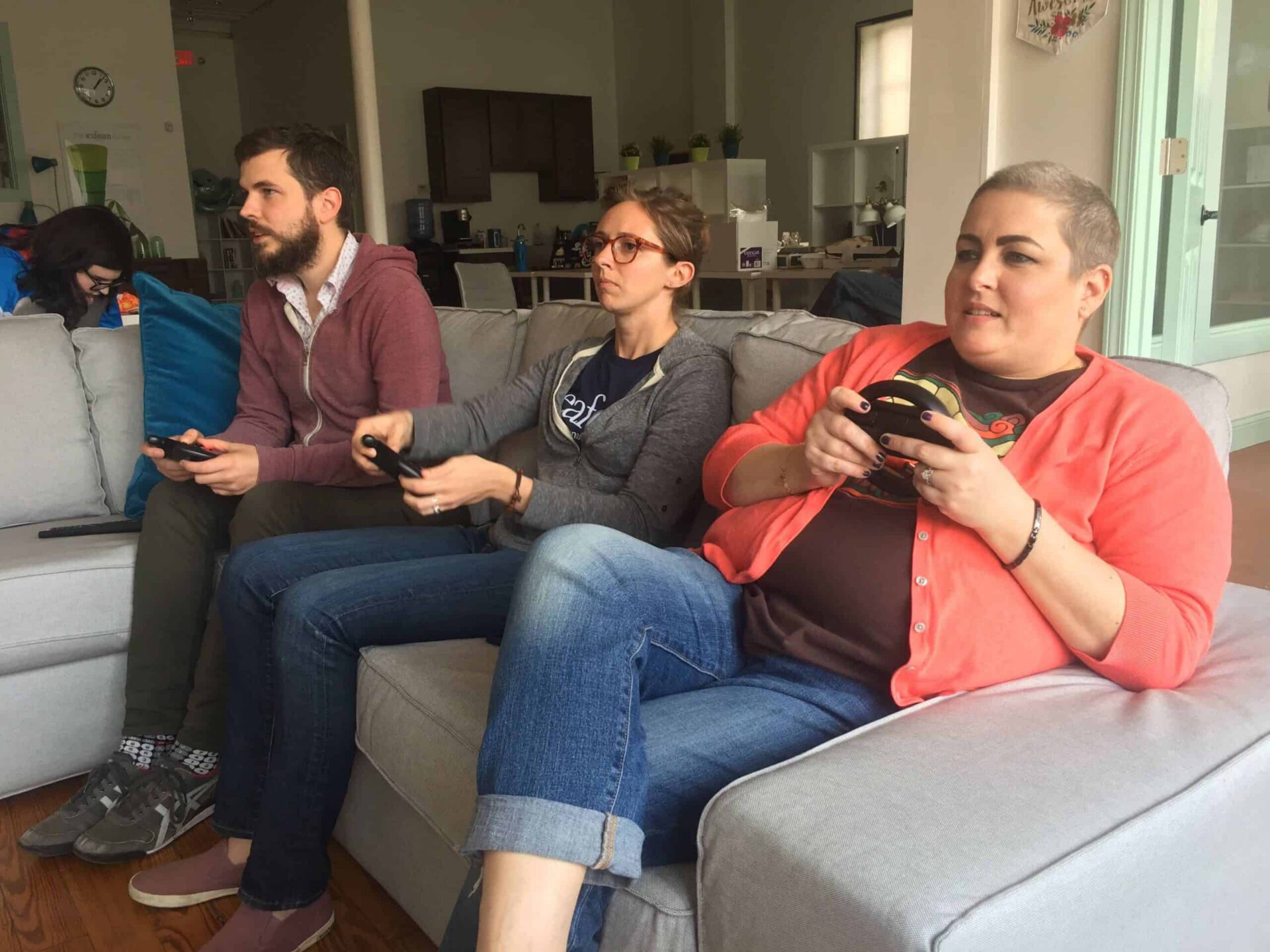 Three adults sitting on a couch and playing video games, each holding a controller, in a cozy living room with a relaxed and focused atmosphere, illuminated by the gentle glow of seafoam day light.