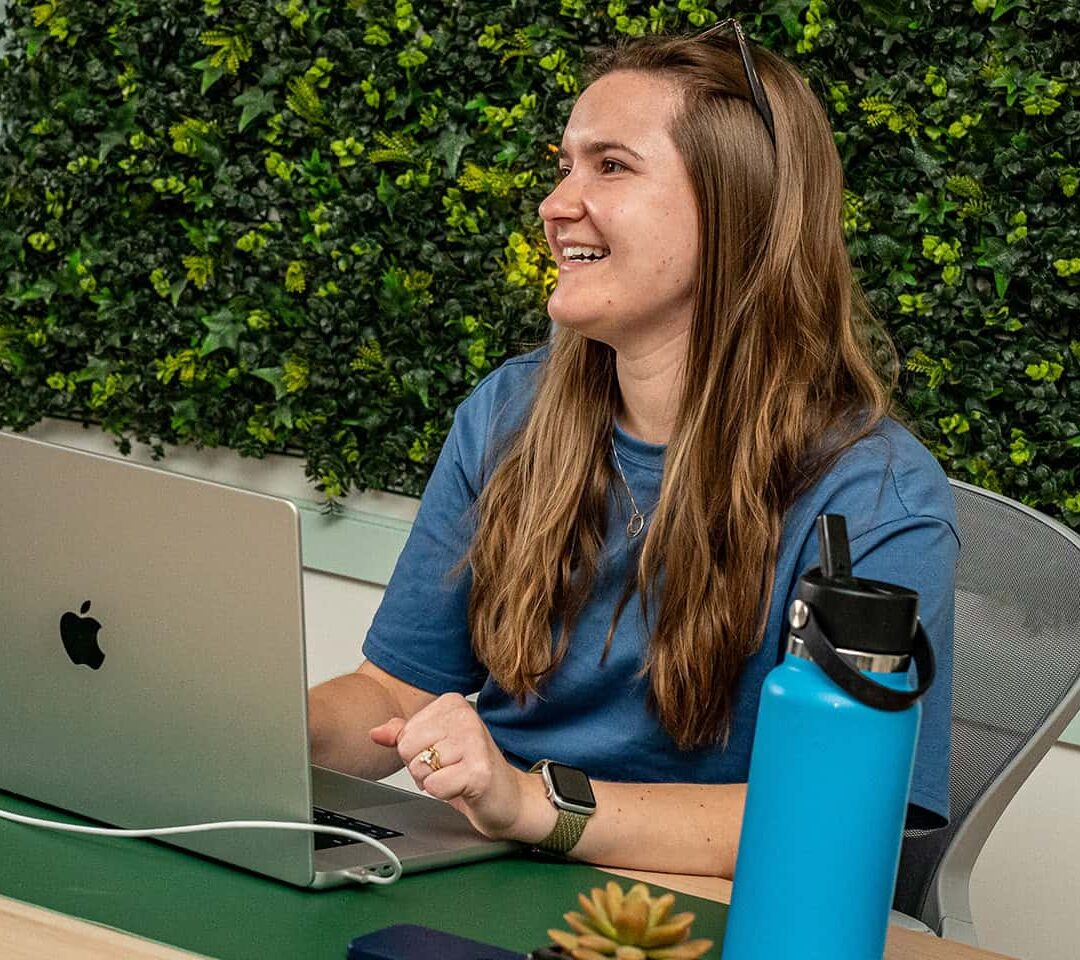 A woman with long hair, smiling, sits at a desk with a laptop, a blue water bottle, and a small potted plant, against a backdrop of a green leafy wall.