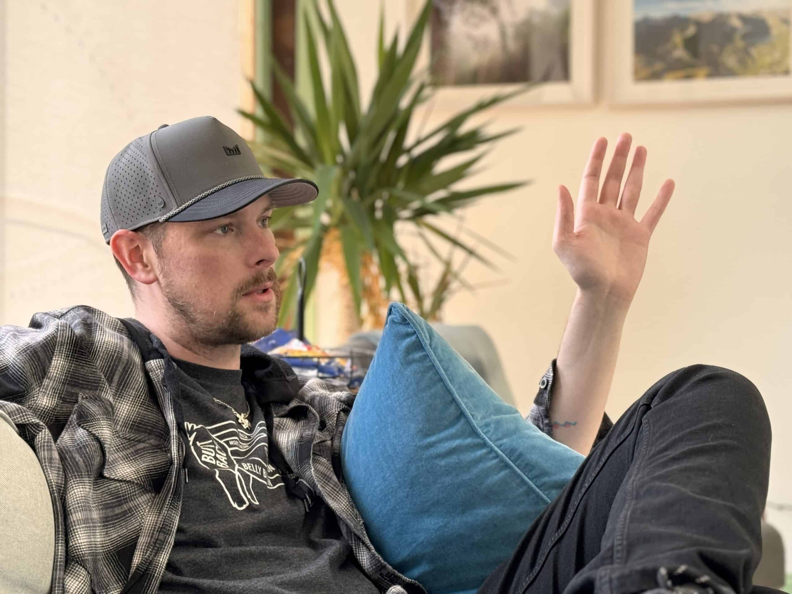 A man with a gray cap and a plaid shirt is seated on a couch, raising his left hand as he speaks. Behind him is a plant and framed pictures on the wall, adding to the cozy indoor setting. He appears to be engaged in a conversation or explaining something.