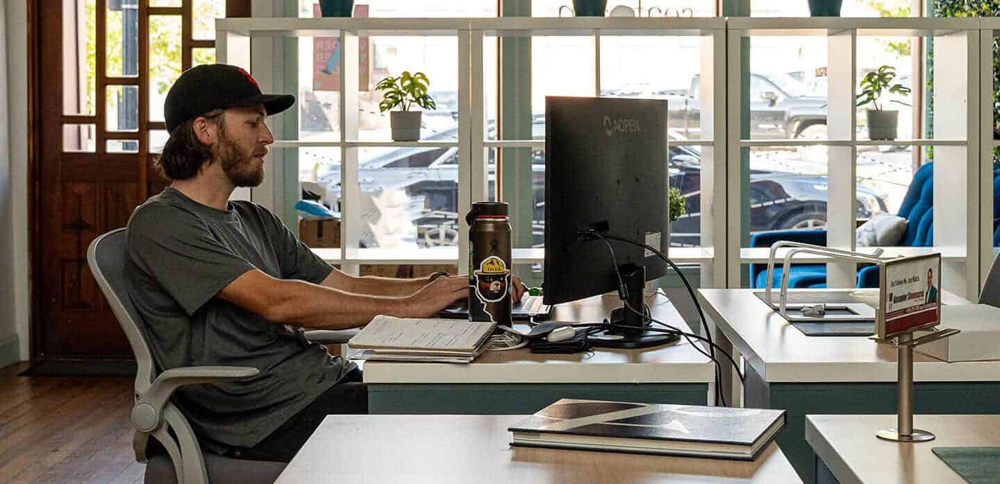 A person wearing a black cap and a gray t-shirt is sitting in front of a computer at a desk in a well-lit office. They are focused on the screen, preparing for Google Marketing Live 2024, and a large coffee mug is placed beside the keyboard. The background shows bookshelves and a street view through windows.