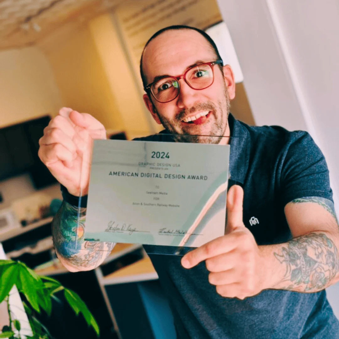 A smiling man with glasses, wearing a gray polo shirt, is holding up a clear plaque that reads "2024 American Digital Design Award." He stands in an indoor setting with a kitchen in the background and a green plant visible in the foreground.