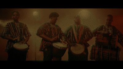 Four people are playing traditional African drums on a dimly lit stage. They are dressed in colorful, patterned shirts and are performing with intensity and focus. The background has a warm, hazy atmosphere with soft spotlights.
