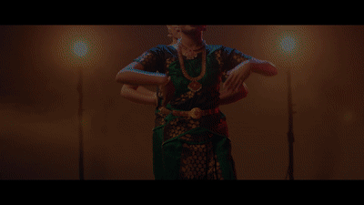 Dancers perform a traditional dance in colorful attire, using expressive hand movements and body postures. The background is dimly lit with warm lights, creating a dramatic effect. The dancers wear intricate costumes and jewelry, adding to the cultural ambiance.