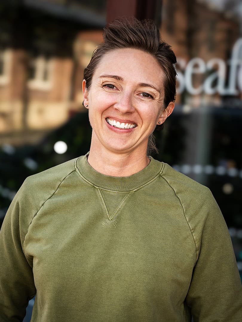 A person with short brown hair is smiling. They are wearing a green sweatshirt and standing in front of a blurred background that includes a glass door or window with partial lettering visible.