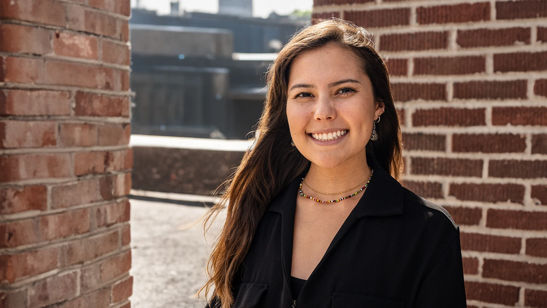 Cenna Mawhinney, with her long brown hair, smiles at the camera. She is wearing a black top, a layered necklace, and small hoop earrings. Standing outdoors between two brick walls, sunlight illuminates her face while the background showcases more brick architecture.