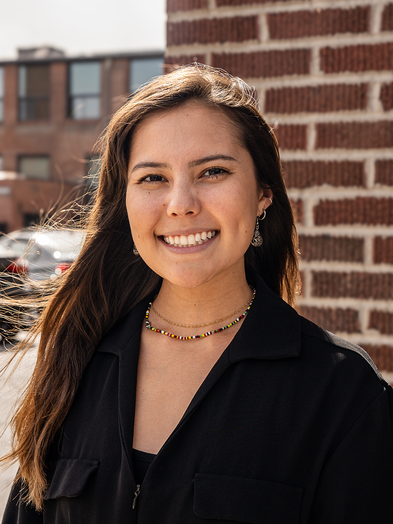 A young woman with long brown hair smiles at the camera, standing outside in front of a brick wall. She is wearing a black top, colorful bead necklace, and earrings. It is a sunny day, and her hair is slightly tousled by the wind.