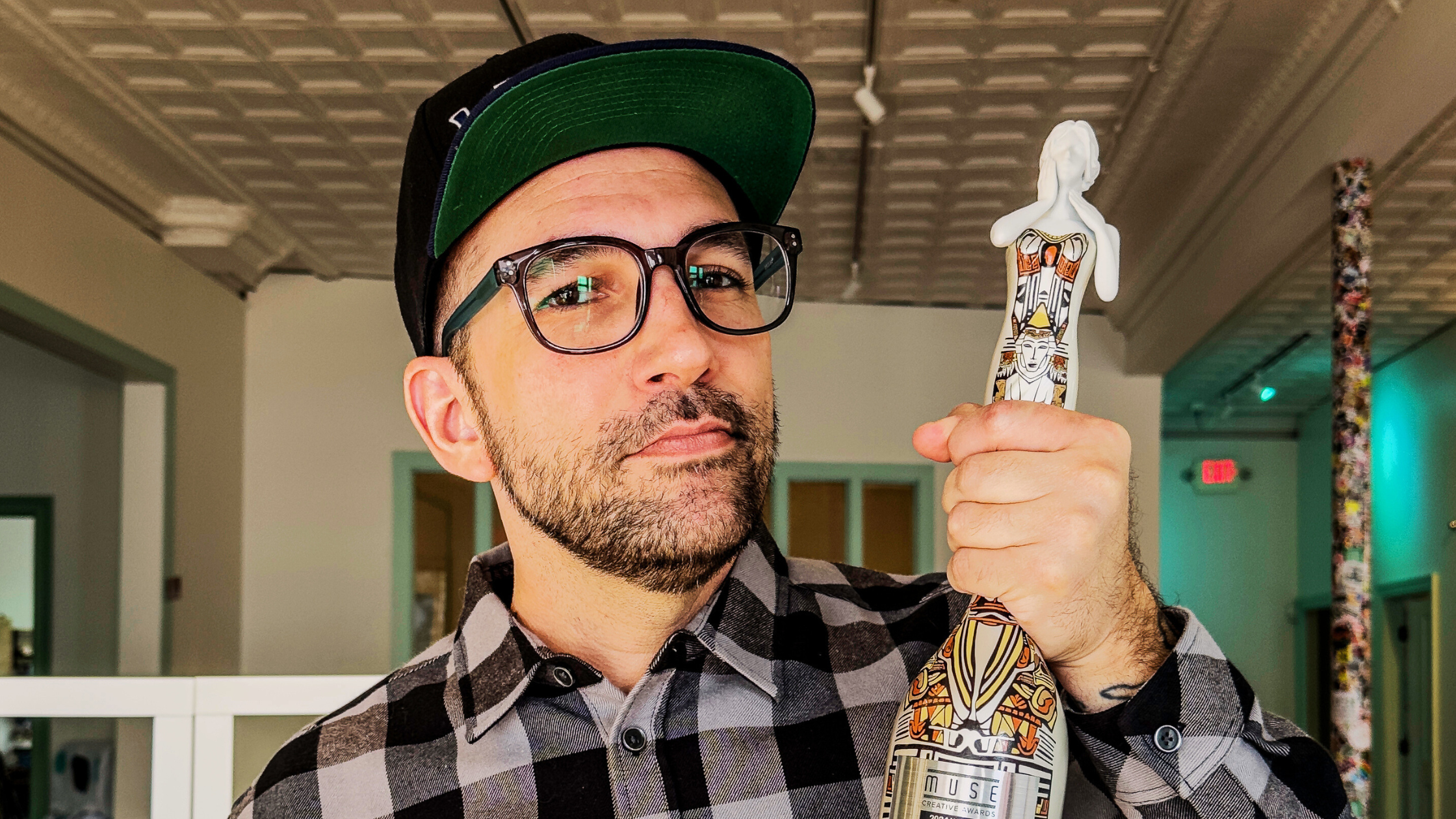 A bearded man wearing glasses and a black cap holds a stylishly decorated bottle with a unique design. Dressed in a black-and-white checkered shirt, he stands indoors against a backdrop of a patterned ceiling with recessed lights, celebrating the Seafoam Wins MUSE Creative Award.