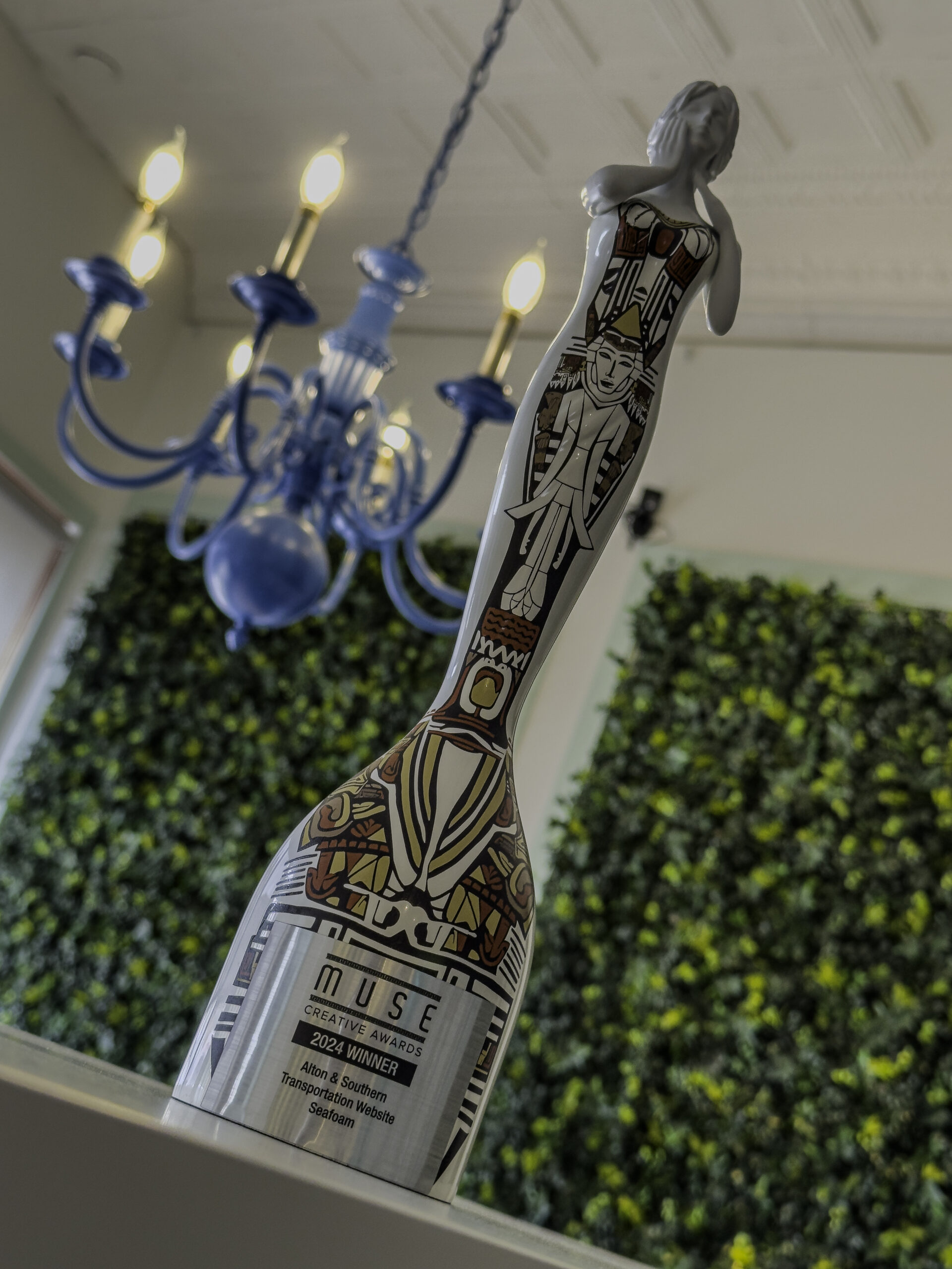 A decorative bottle featuring intricate, artistic designs stands on a table. Above it is a blue chandelier with lit bulbs. In the background, there's a wall covered in green foliage and a window letting in natural light. The bottle's label proudly reads "Seafoam Wins MUSE Creative Award.