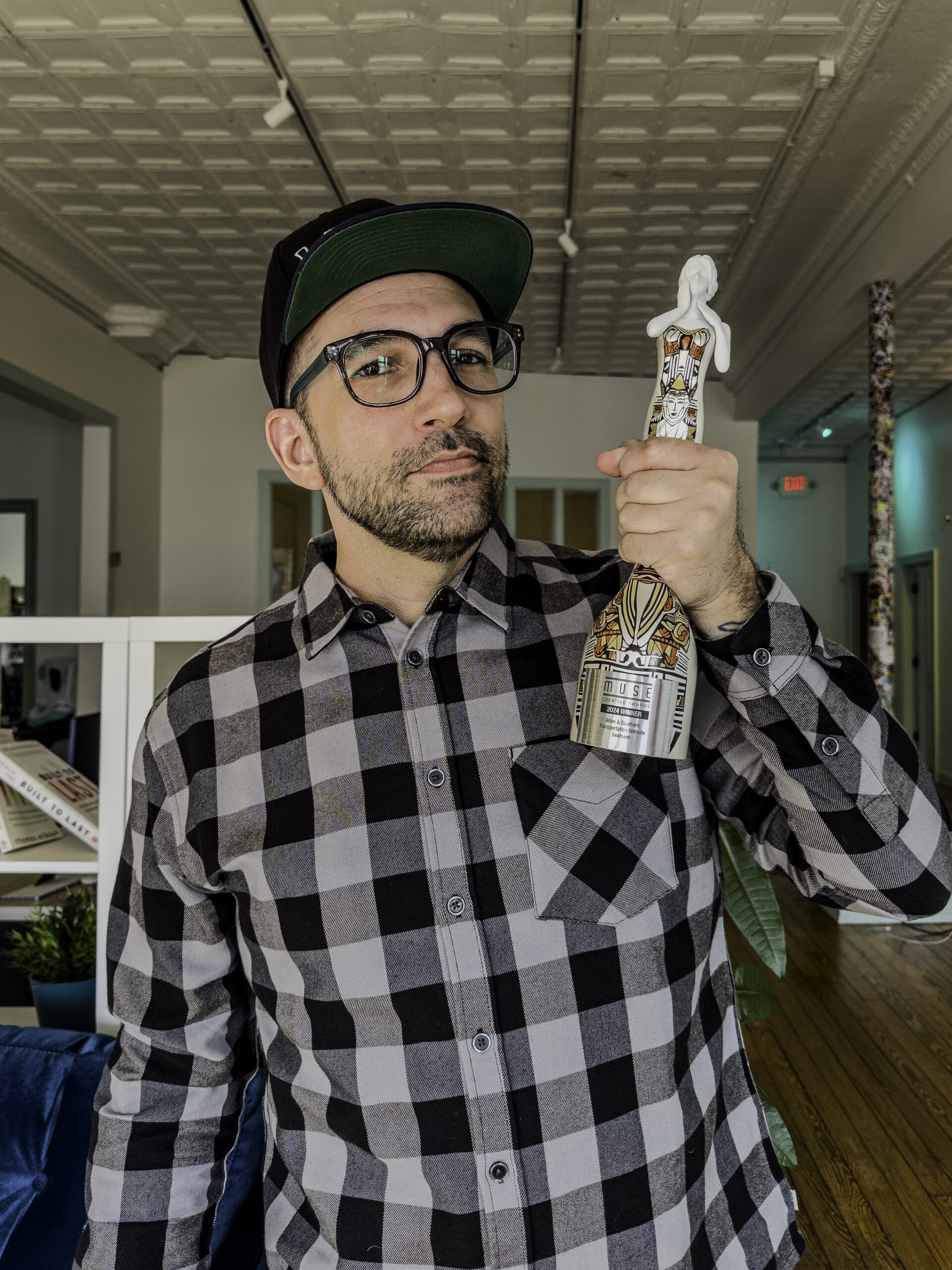 A bearded man in a black and white plaid shirt, wearing glasses and a black cap, proudly holds up a decorative bottle with an intricate pattern and figure on top. He's standing in a room with a tiled ceiling and wooden floor, celebrating as Seafoam wins the MUSE Creative Award.