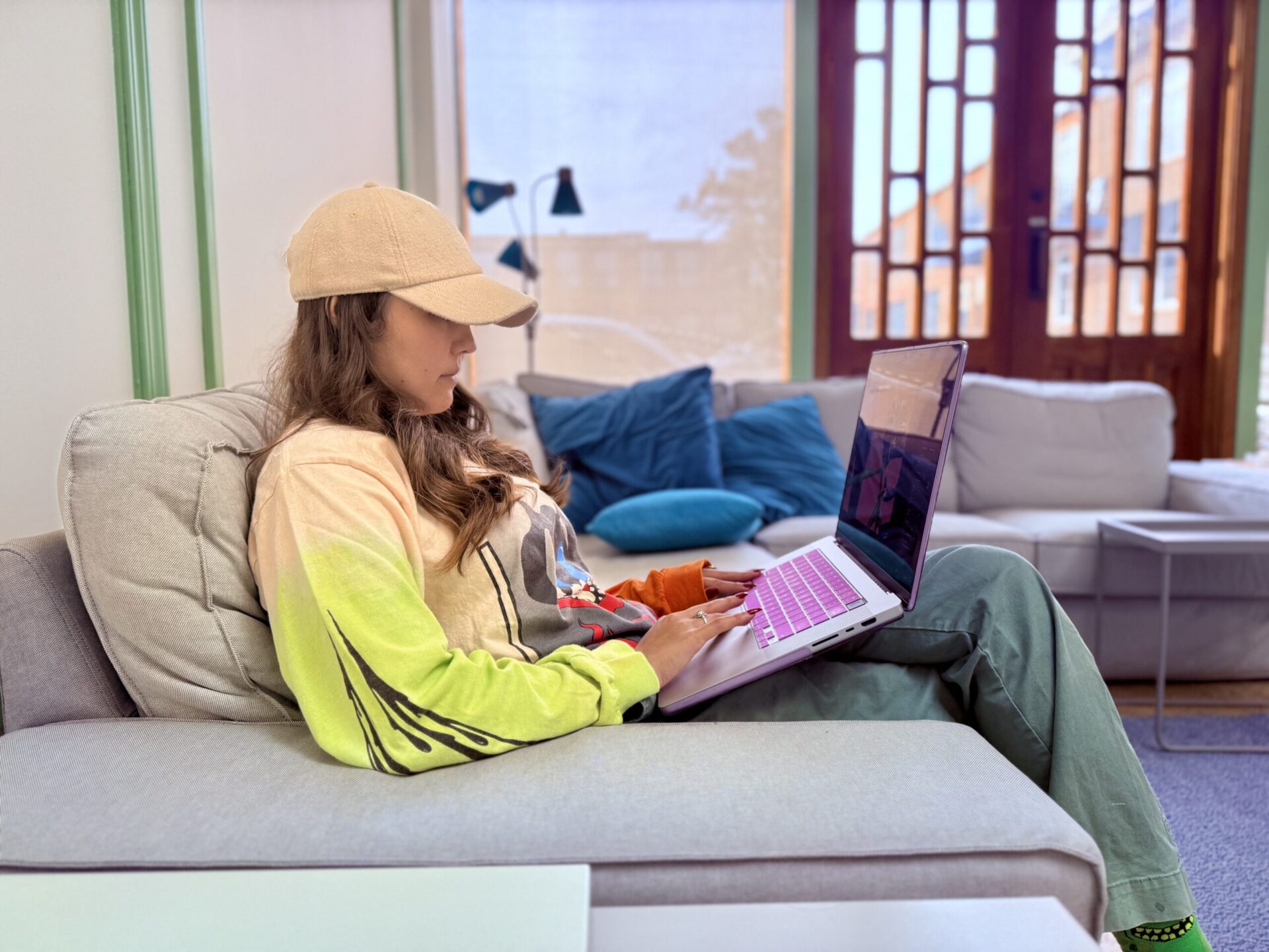 A person wearing a beige cap and colorful hoodie is sitting on a light gray sofa, using a laptop. The room has large windows, blue cushions, and a bright, airy feel.