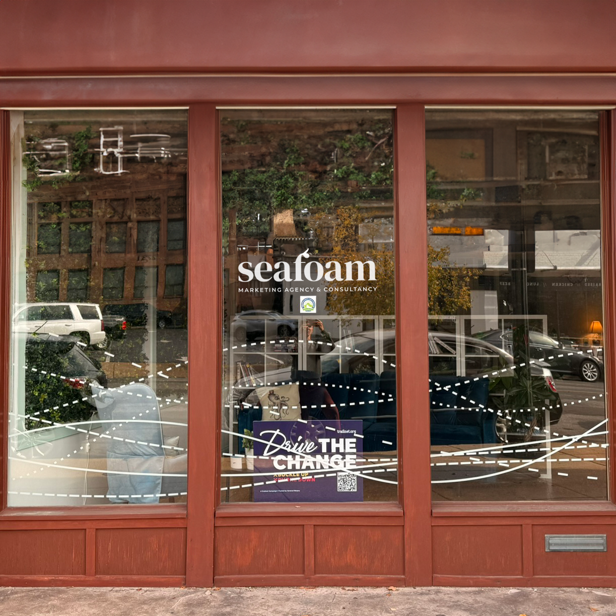 Storefront of Seafoam Marketing Agency & Consultancy with large windows and a red frame. The window displays the slogan "Drive the Change" with street reflections visible.