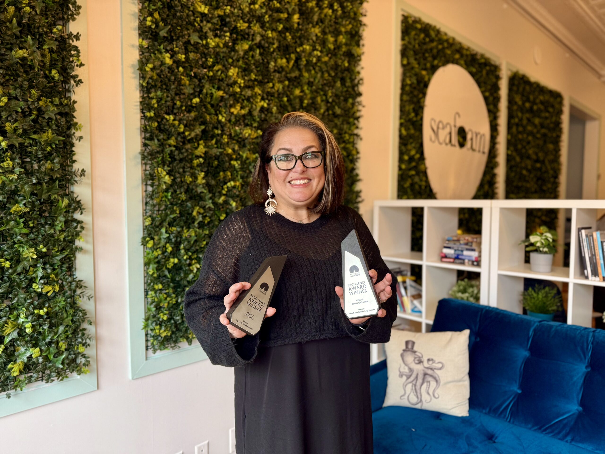 A person wearing glasses holds two awards while smiling. They stand in front of a wall adorned with green plants and a logo that reads "seafoam." A shelf with books and a blue couch are in the background.