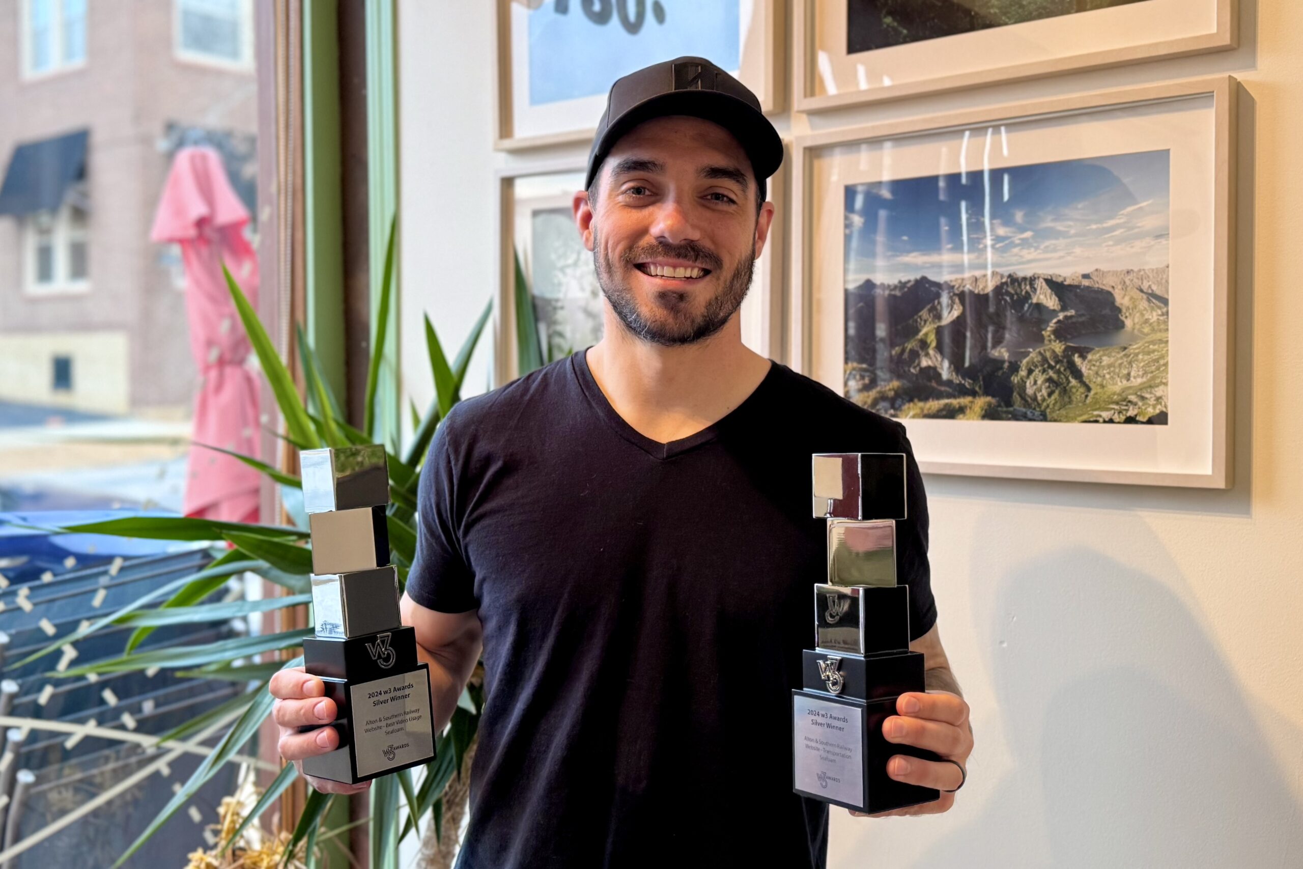 A man in a black T-shirt and cap is smiling and holding two awards indoors. The background features framed pictures on the wall and a window with a view of a red umbrella outside.