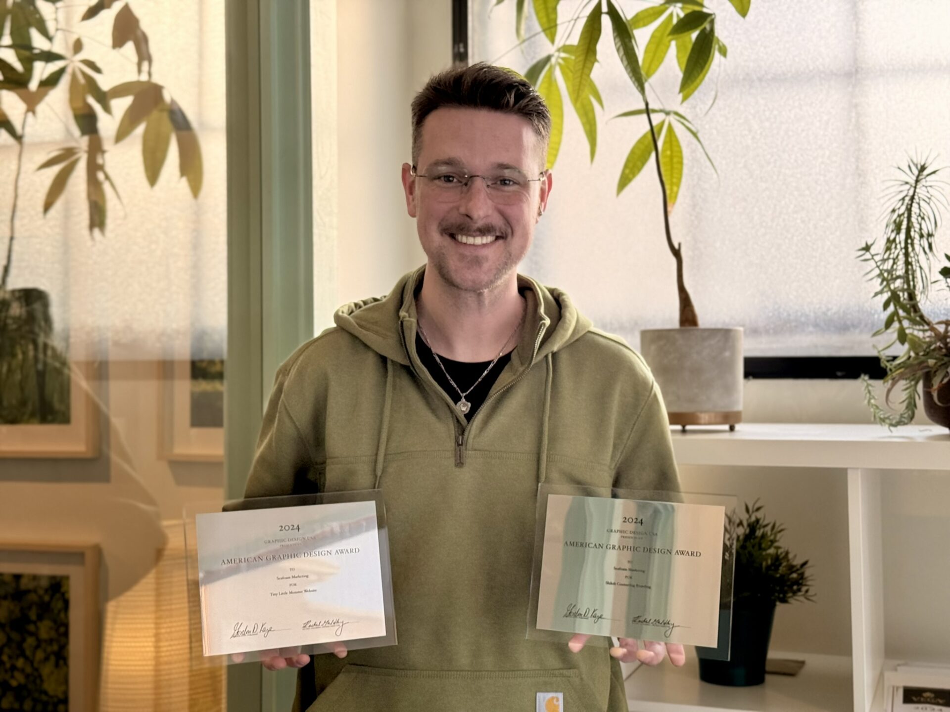 A person wearing a green hoodie and glasses smiles while holding two awards labeled "American Legion Citation Award" in front of a shelf with plants and framed pictures.