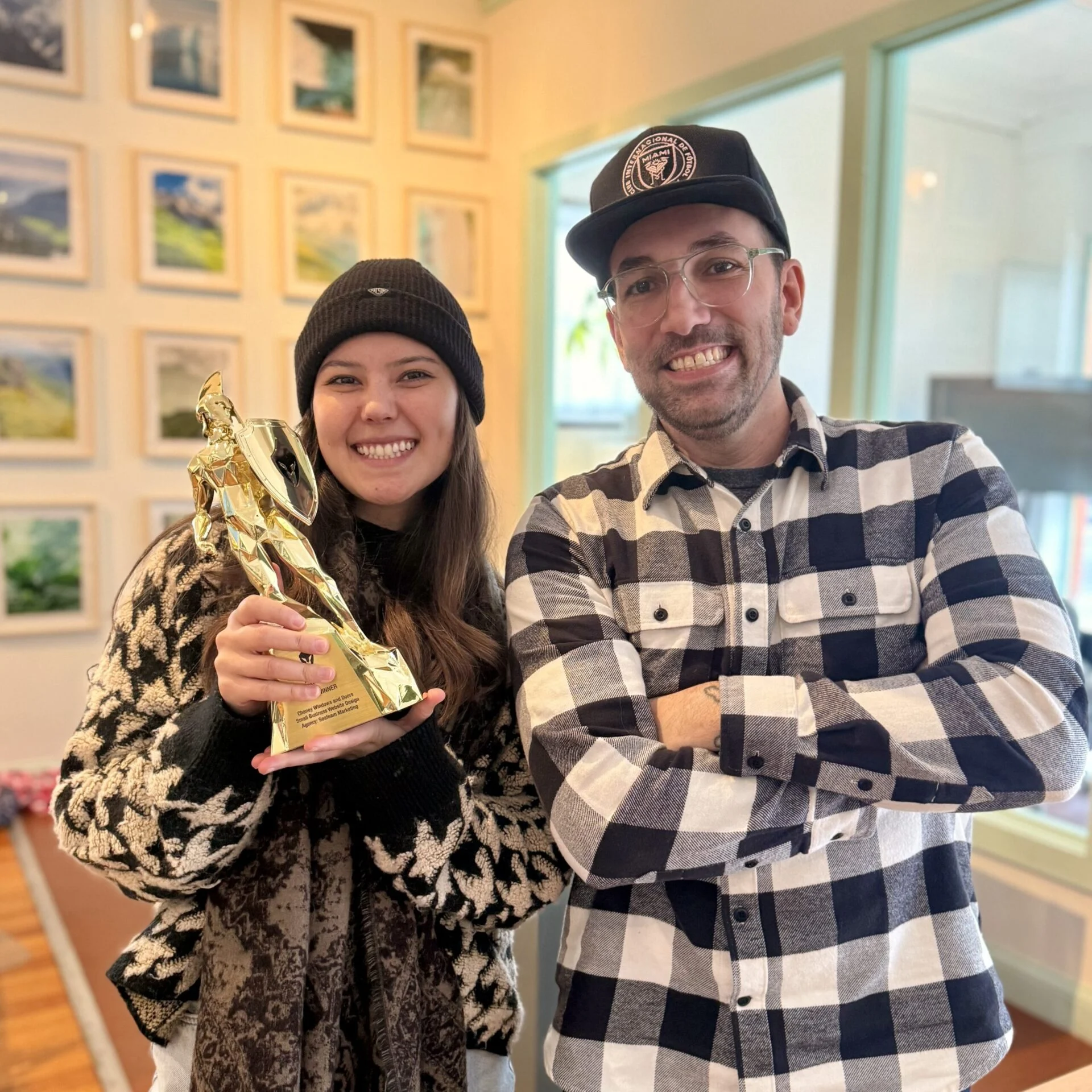 A smiling woman in a beanie and scarf holds a gold trophy while standing next to a man in glasses and a cap, wearing a plaid shirt. They are indoors with framed pictures on the wall behind them.