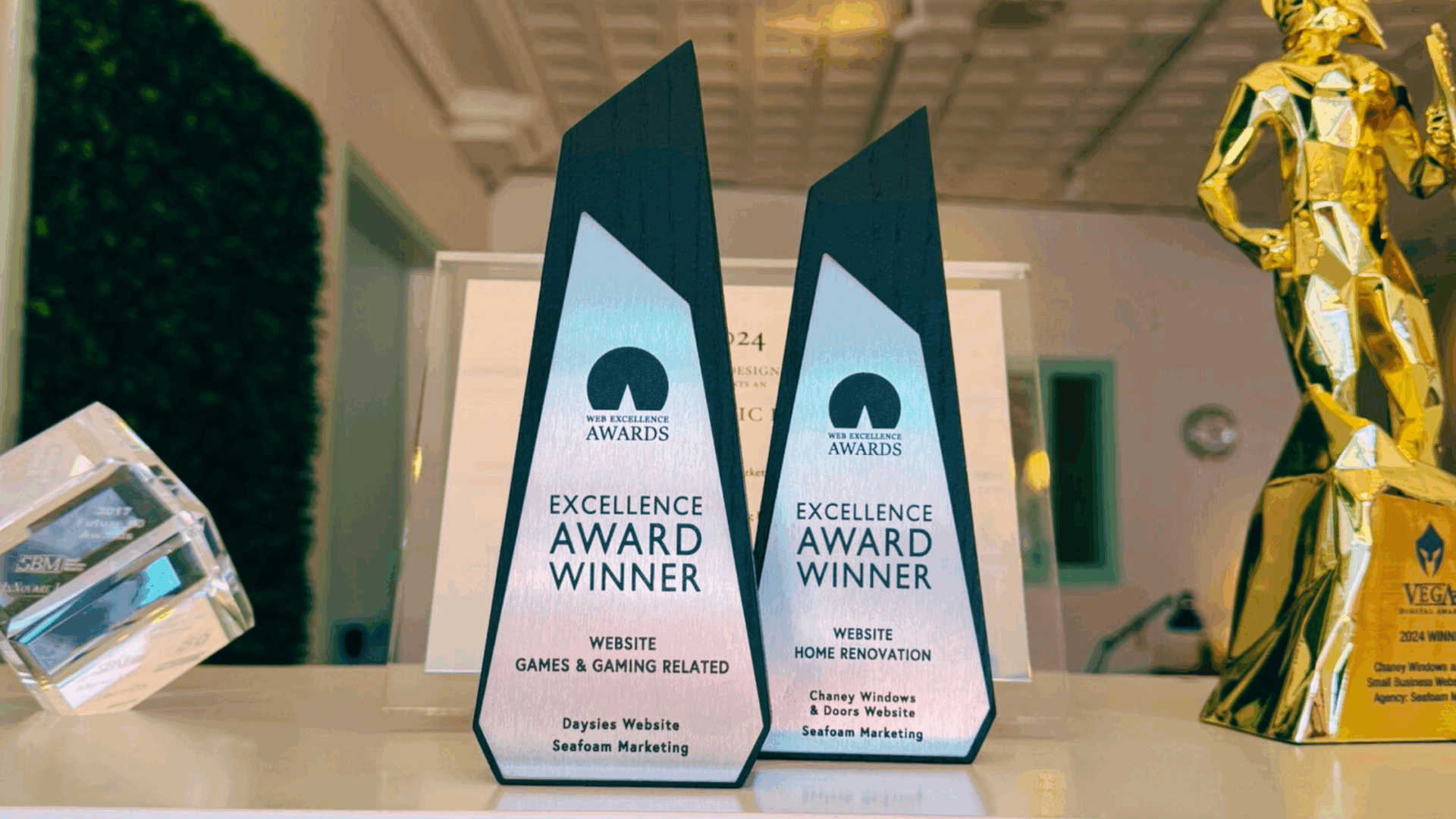 Two black and silver award trophies labeled "Excellence Award Winner" for website design stand on a white surface. One is for "Games & Gaming Related" and the other for "Home Renovation." A blurred room is in the background.