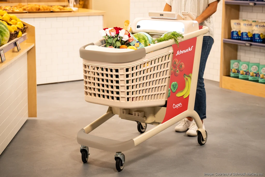A person pushes a shopping cart filled with groceries, including fresh produce and flowers, inside a store. The cart, showcasing innovative St. Louis marketing, features a digital screen and a red panel adorned with banana illustrations. Shelves of fruits are visible in the background.
