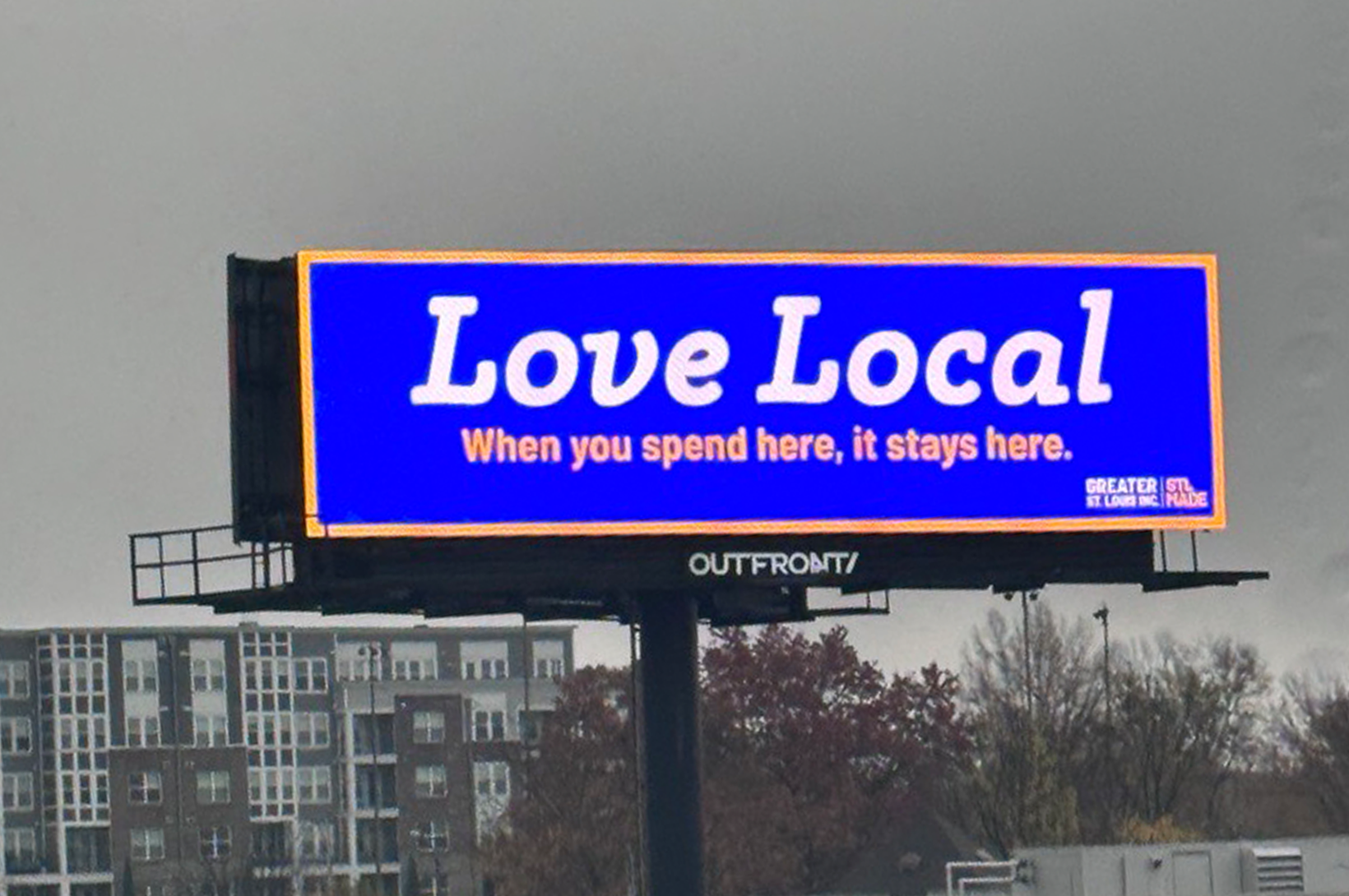 A billboard in St. Louis reads "Love Local" in large white text on a blue background, with smaller text stating, "When you spend here, it stays here." Trees and buildings provide an urban backdrop to this clever piece of local marketing.