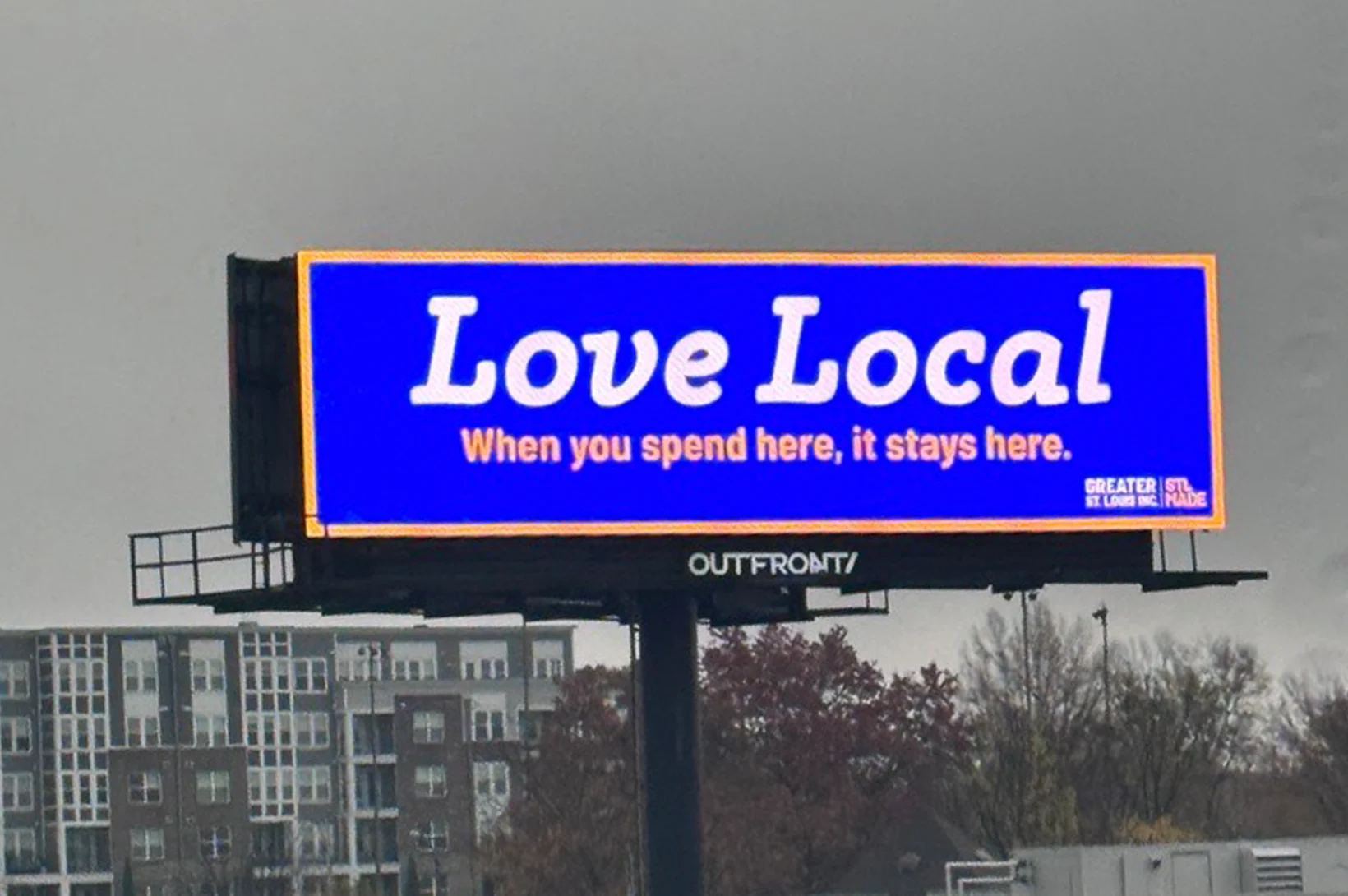 A billboard in St. Louis reads "Love Local" in large white text on a blue background, with smaller text stating, "When you spend here, it stays here." Trees and buildings provide an urban backdrop to this clever piece of local marketing.
