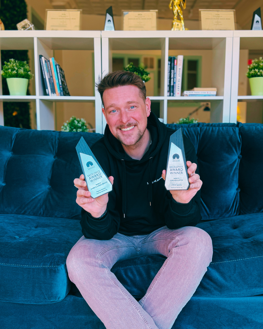 A person sitting on a blue couch holding two award trophies with a proud smile. Behind them is a white bookshelf filled with books and small plants.