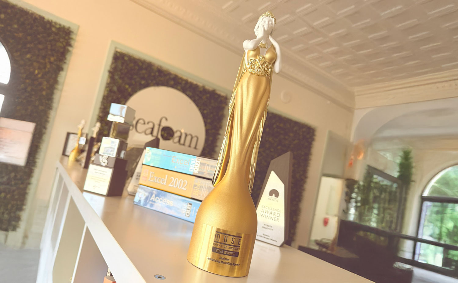 A golden trophy shaped like a woman in a gown is displayed on a shelf, celebrating an Outstanding Marketing Agency, surrounded by books and other awards in a well-lit modern office with greenery and arched windows.
