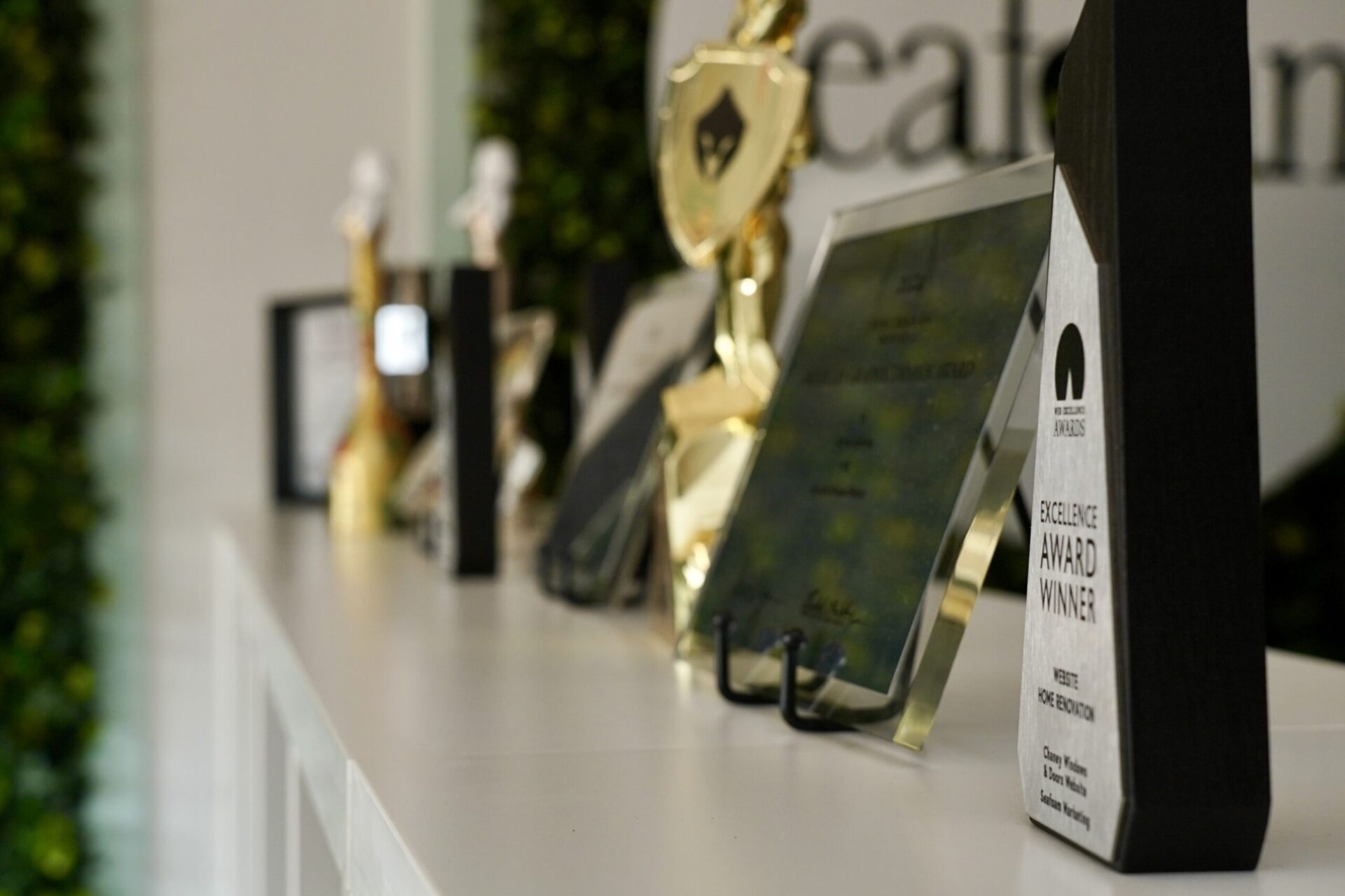A row of various awards and trophies is displayed on a white shelf, with some plaques and golden statues in focus. The background is blurred with greenery and part of a logo visible.