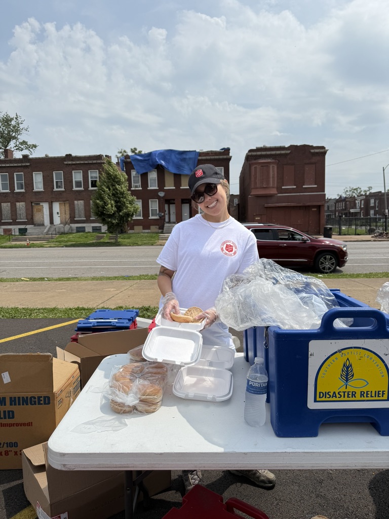 A smiling volunteer in a white shirt and cap prepares sandwiches at an outdoor disaster relief station, with food containers and supplies on a table in front of them and old brick buildings in the background.