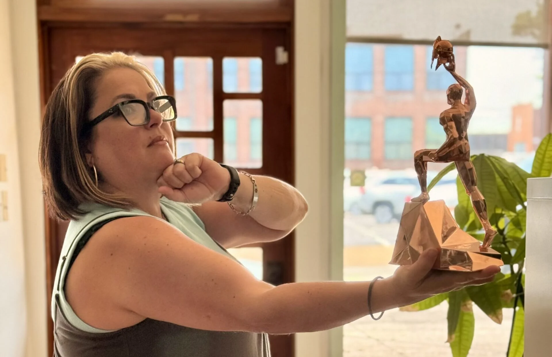 A woman wearing glasses holds a bronze sculpture of a person in a thinking pose, mimicking the pose herself while standing indoors near a potted plant with sunlight streaming through the window behind her.
